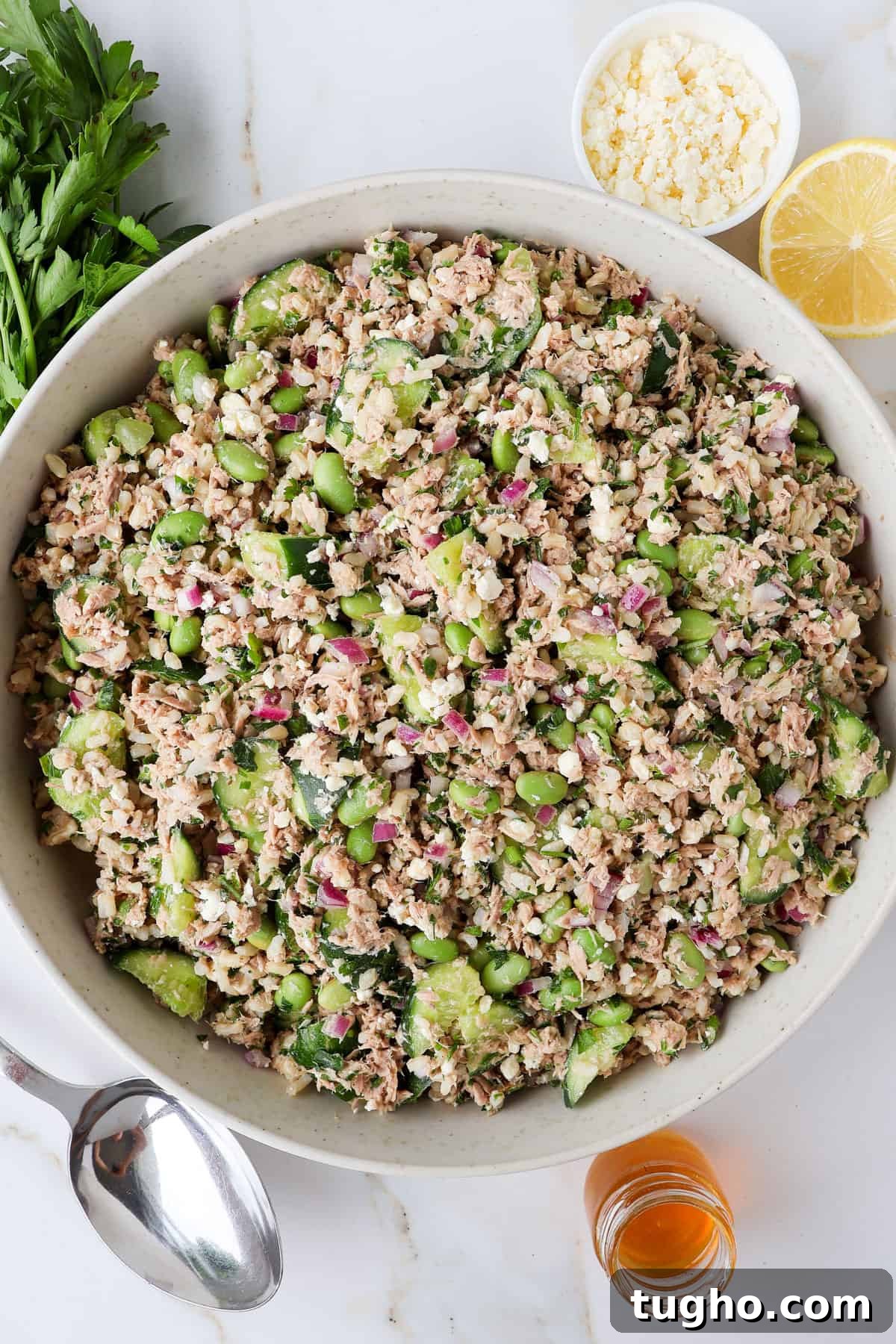 Protein-Packed Tuna, Cucumber and Edamame Salad Recipe 2 Tuna cucumber salad in a bowl with parsley, feta, lemon and honey in a jar on the sides.
