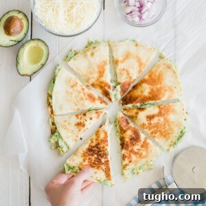 30 Minute Dinner Recipes for Busy Weeknights 13 A hand takes a slice of a quesadilla on a white surface. The quesadilla is cut into eight pieces and filled with avocado. Nearby are bowls of shredded cheese and chopped red onion, a half avocado, and a pizza cutter beside a blue-checkered cloth.
