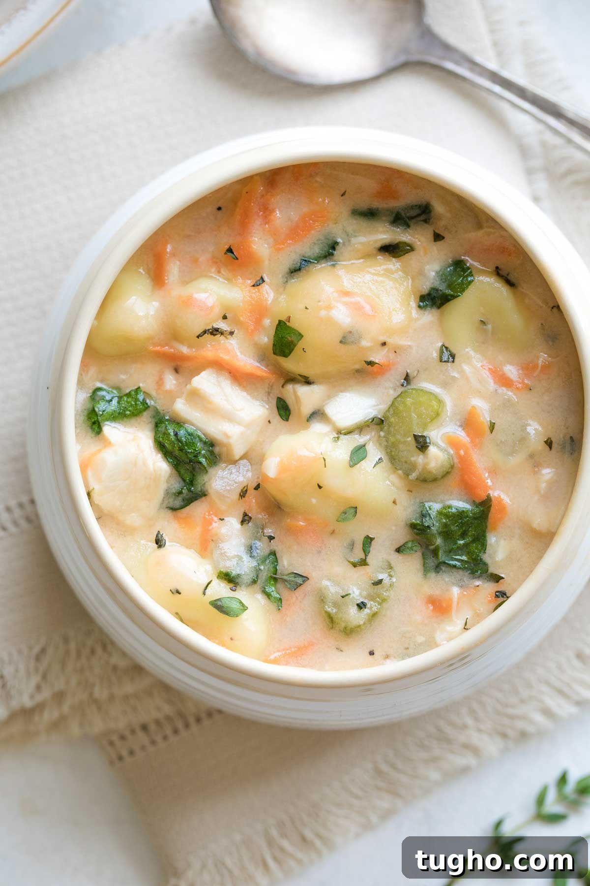 Overhead of a white bowl with creamy colored rim on cream napkin, with spoon above and fresh thyme sprigs at corner.