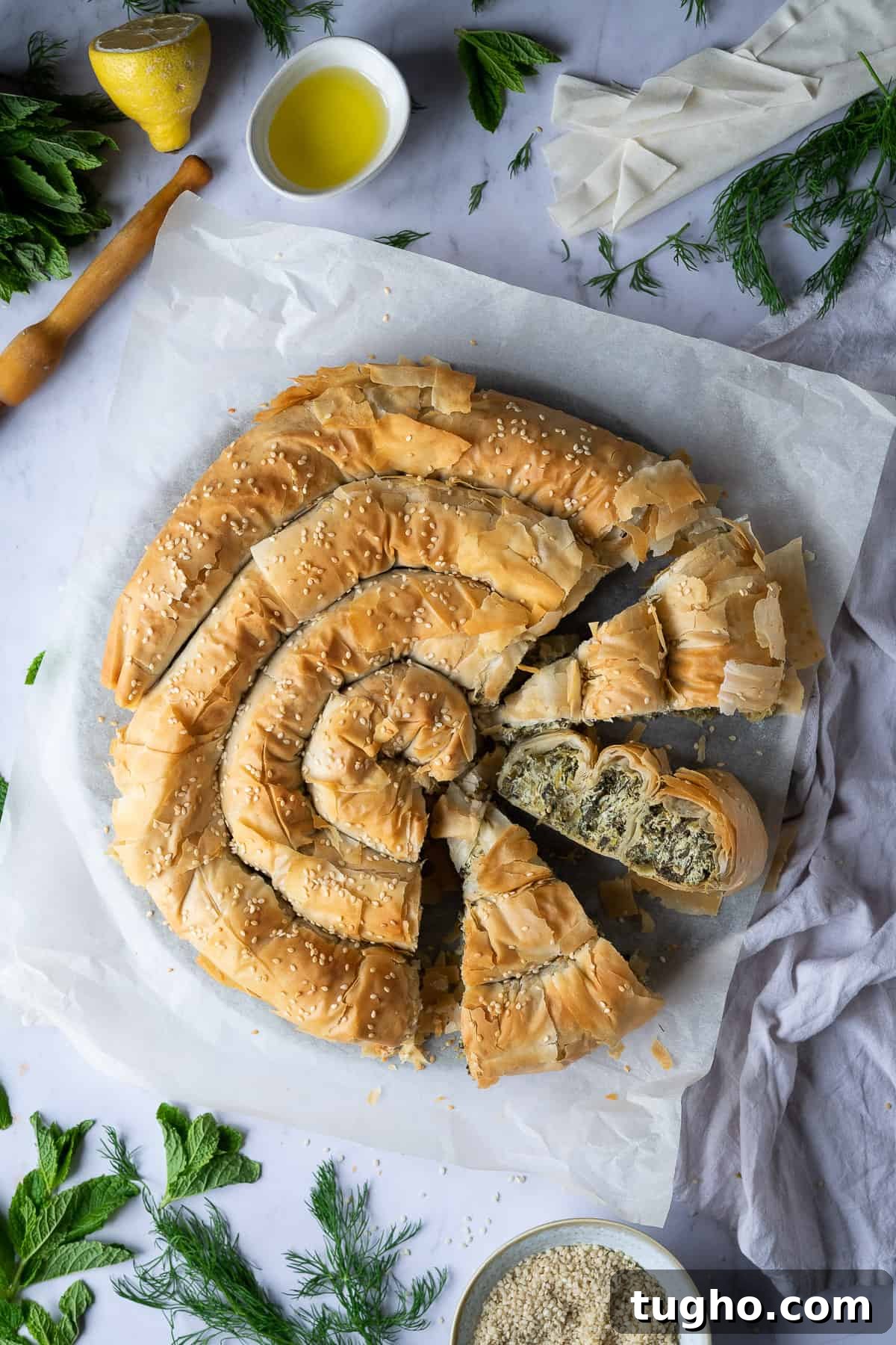 Vegan spanakopita spiral - this impressive vegan spinach and cheese filo pie consists of a soft, cheesy, salty, herby spinach filling encased in crisp phyllo pastry. It's a real crowd pleaser that is much easier to make than it looks! #veganbaking #dairyfree #veganmeal Sliced vegan spanakopita spiral on a sheet of baking parchment on a marble background with fresh herbs and olive oil.