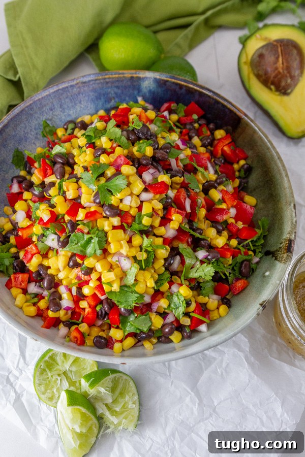 looking down into a bowl of corn salad, served with lime wedges, avocado, and dressing.