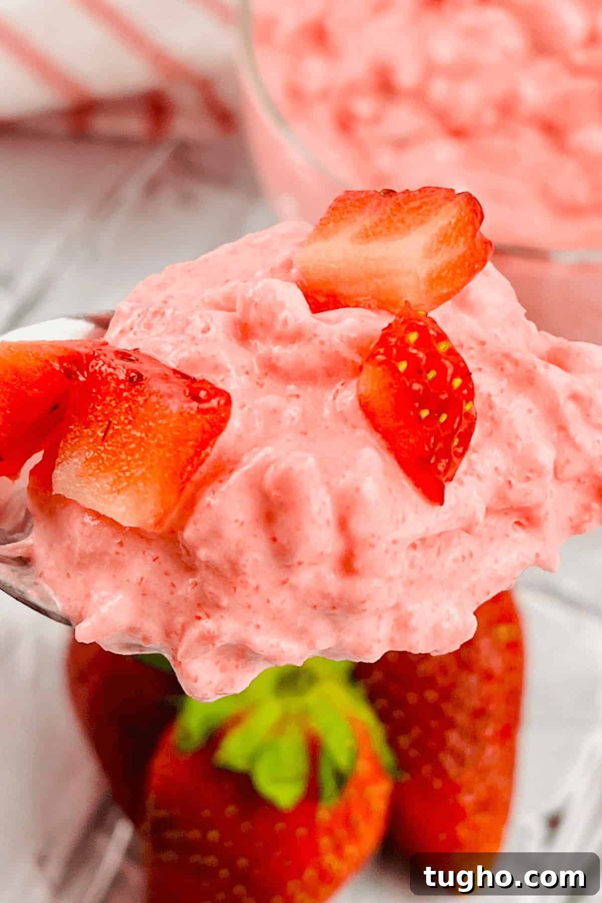 A spoonful of pink strawberry mousse reminiscent of a classic cottage cheese jello salad, topped with small strawberry pieces, with whole strawberries in the background.