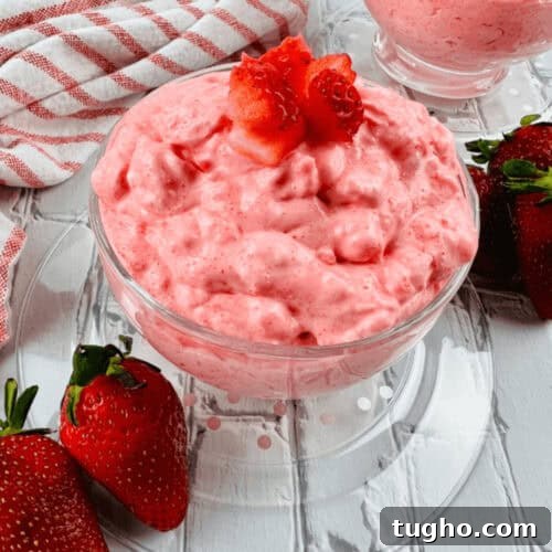 Two glass bowls of creamy pink strawberry dessert, reminiscent of a Cottage Cheese Jello Salad, are topped with strawberry pieces. They sit on a white wooden surface with whole strawberries and a striped cloth nearby.