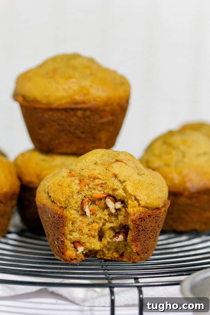 Moist Gluten-Free Carrot Muffins 3 Image shows a close up of gluten free carrot muffins on a cooling rack, with one stacked on another in the background. Front muffin has a small bite taken out of it, showing pecans inside.