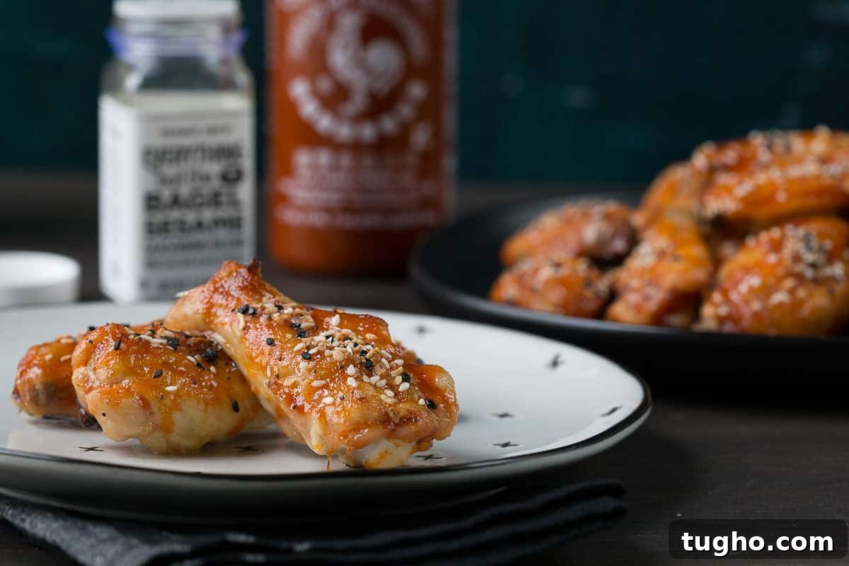 Oven-Baked Sweet and Spicy Chicken Wings Recipe 3 baked chicken wings on a white plate next to a bottle of sriracha and a jar of everything bagel seasoning.