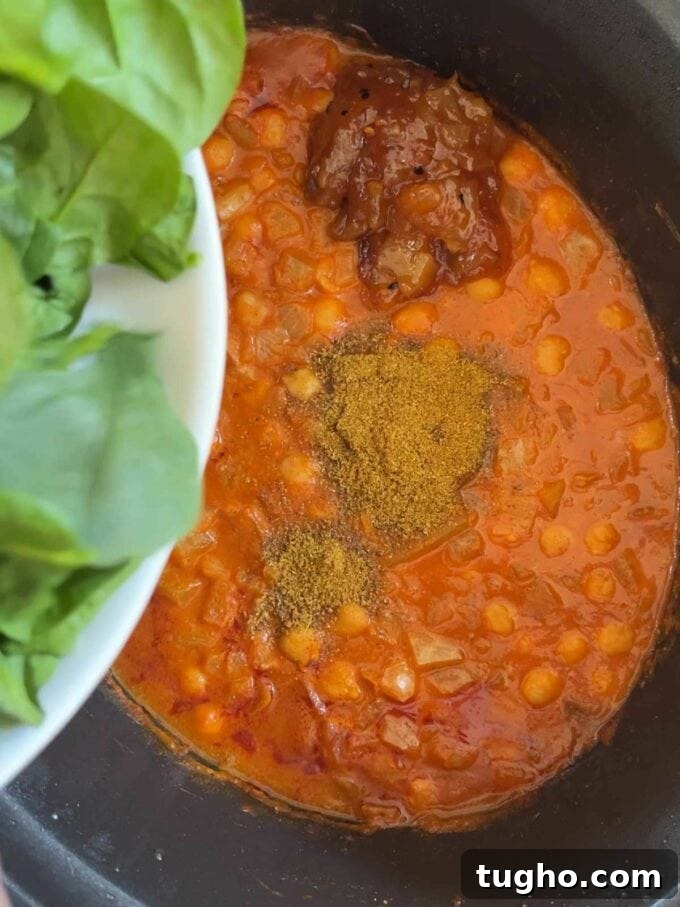 Slow Cooker Chana Saag: Chickpea Curry with Spinach 6 A slow cooker pan filled with chickpeas in a sauce, with spices on top and spinach about to the tipped in.