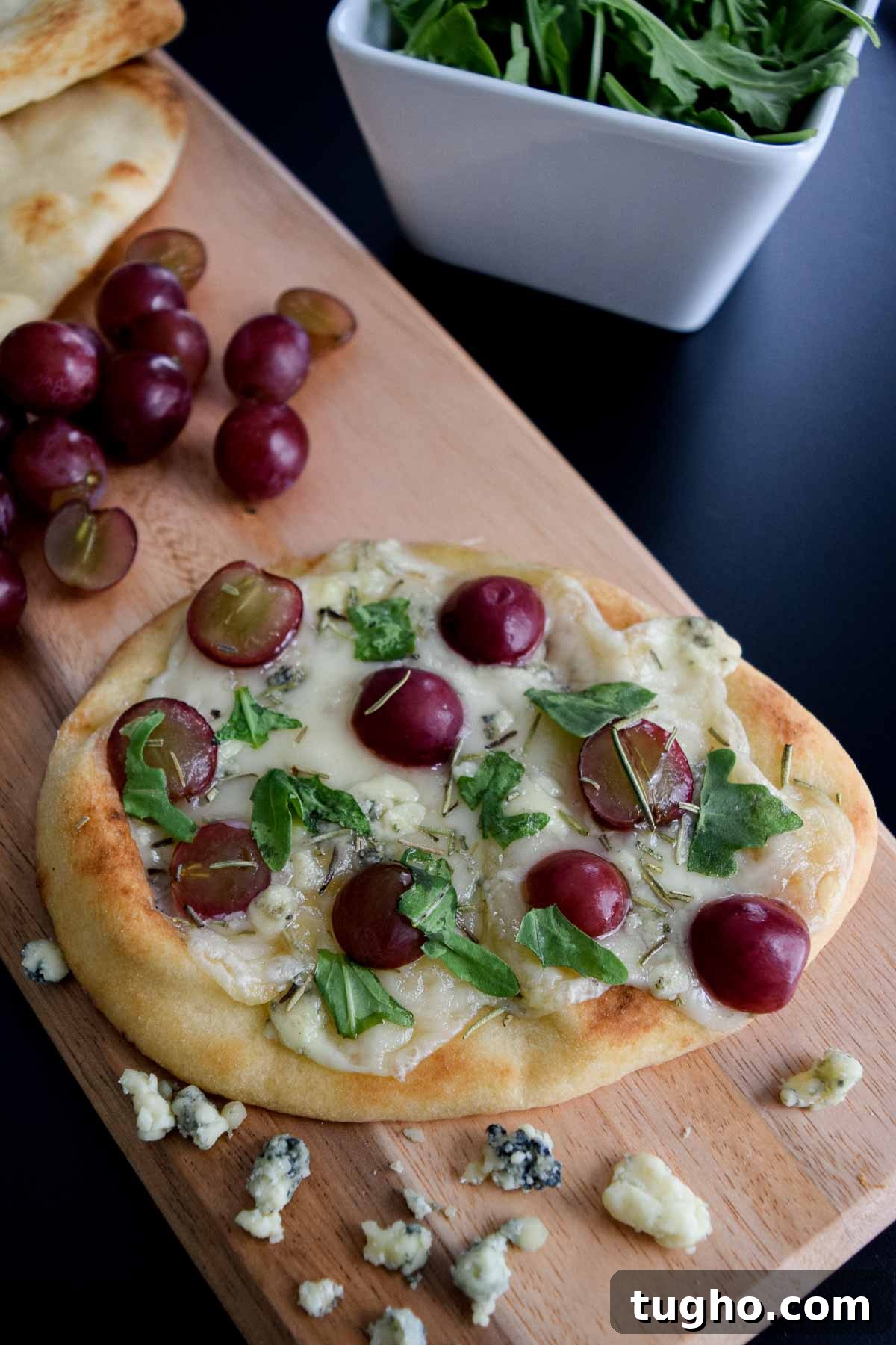 close up view of a flatbread topped with cheese grapes and arugula sitting on a wooden serving board