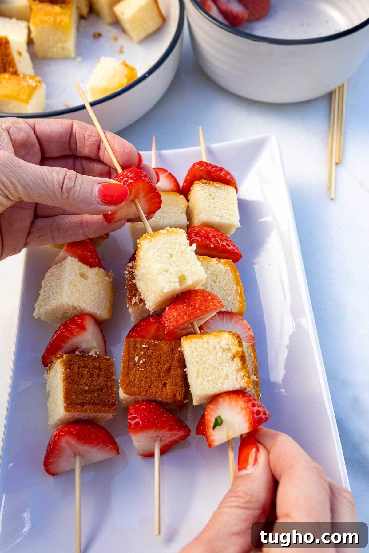 Strawberry Angel Food Cake with Chocolate Sauce 4 Someone's hands threading half strawberries and cubes of cake onto a skewer, with more strawberry skewers below on a plate.