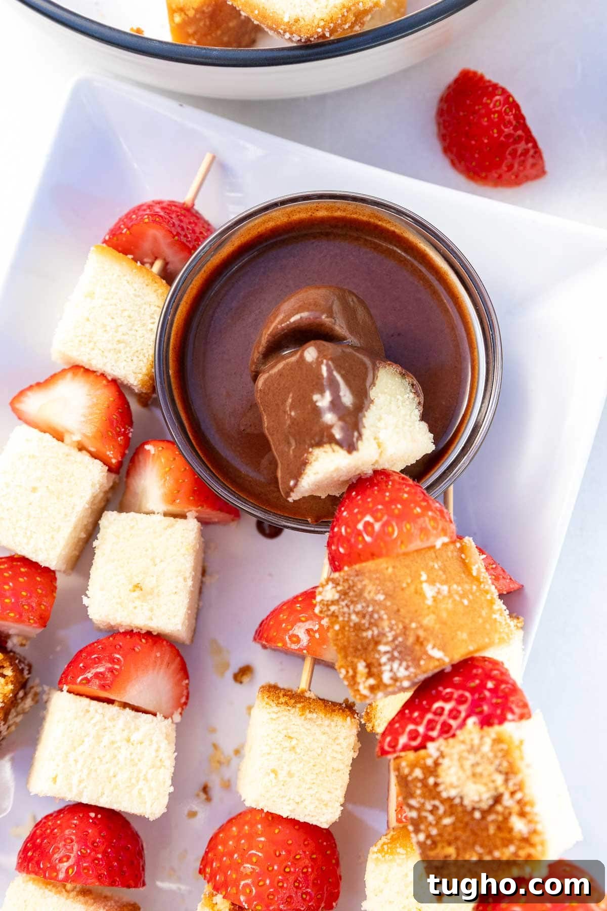 Strawberry Angel Food Cake with Chocolate Sauce 8 Closeup of half strawberries and cubes of cake threaded onto wooden skewers, one with the end dipped into a small bowl of chocolate sauce.