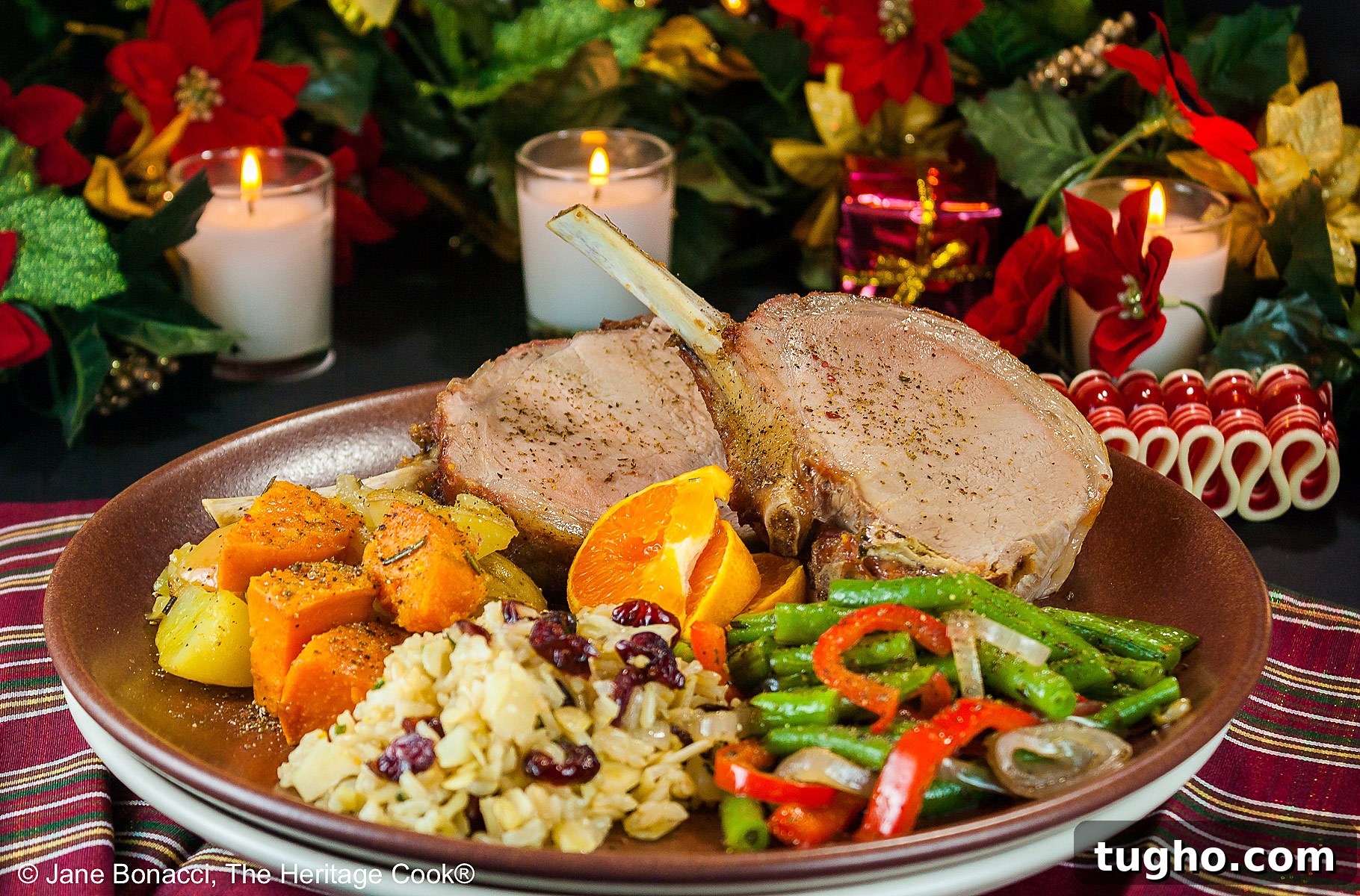 Showstopping Grilled Crown Roast of Pork Recipe 2 Grilled Crown Pork Roast sliced into chops and placed on a dark brown plate with green beans, rice pilaf, and sweet potatoes, surrounded by white candles, green garland with red accents for a festive holiday table © 2024 Jane Bonacci, The Heritage Cook.