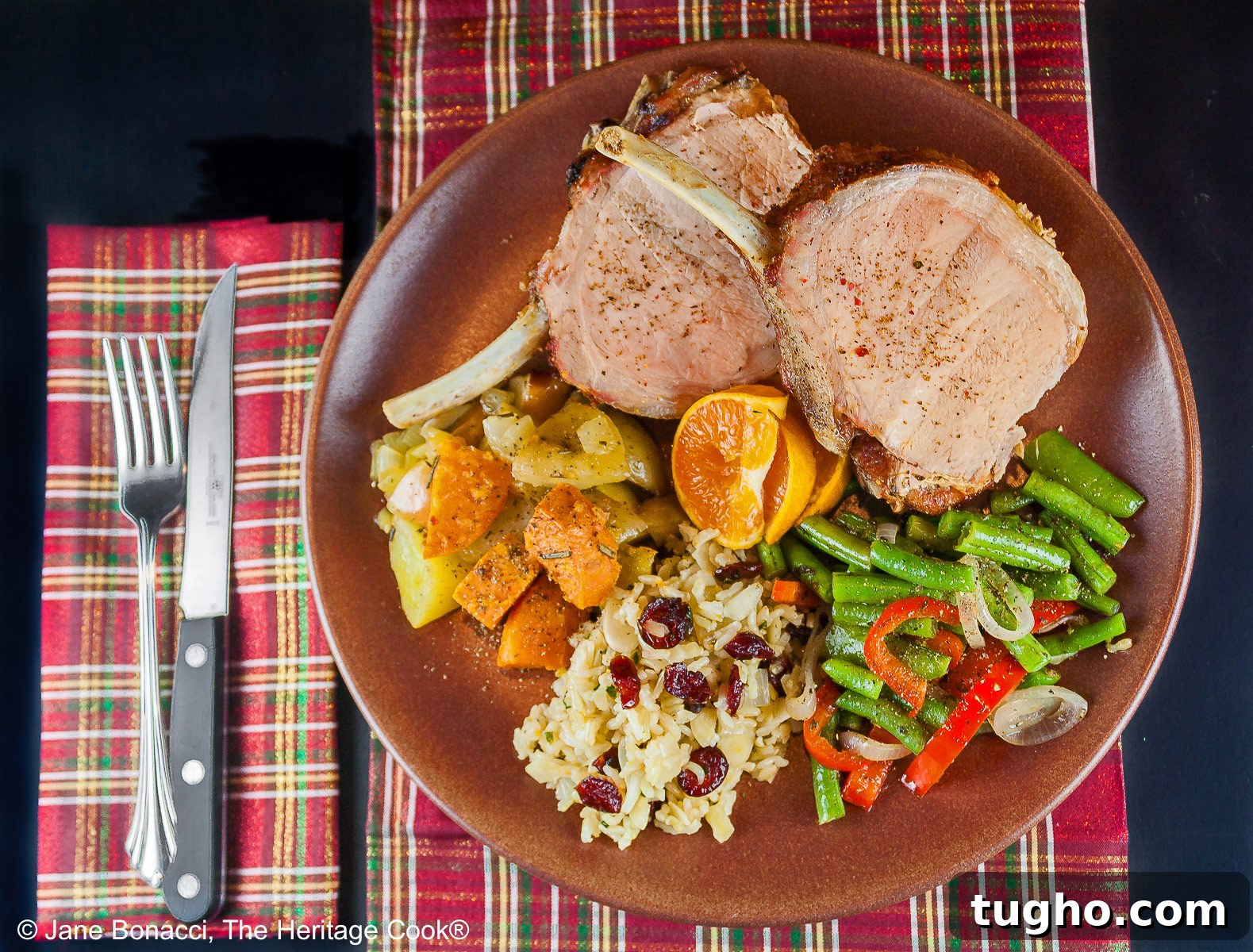 Showstopping Grilled Crown Roast of Pork Recipe 5 Grilled Crown Pork Roast sliced into chops and placed on a dark brown plate with green beans, rice pilaf, and sweet potatoes, surrounded by white candles, green garland with red accents for a festive holiday table © 2024 Jane Bonacci, The Heritage Cook.