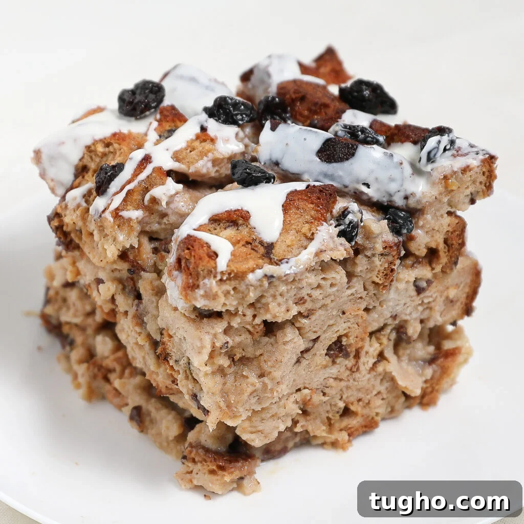 Cinnamon Raisin Bread Pudding Recipe 2 A thick square slice of Cinnamon Raisin Bread Pudding topped with raisins and drizzled with white icing sits on a white plate. The bread is golden brown and appears moist and dense.