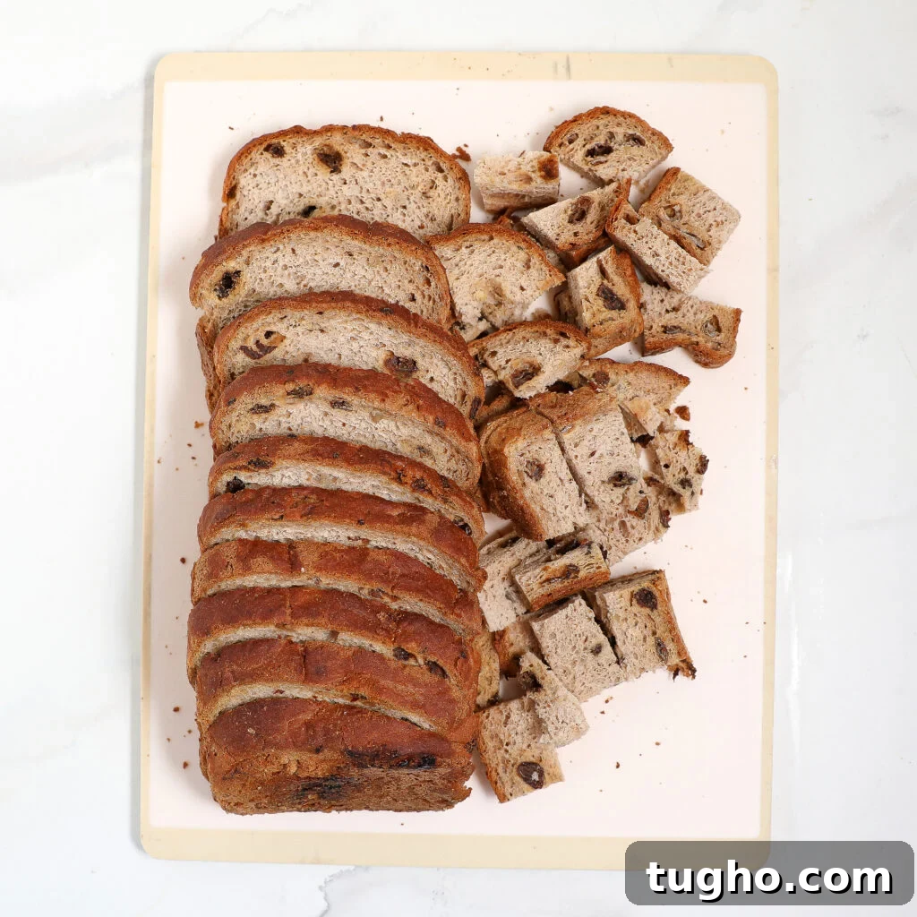 Cinnamon Raisin Bread Pudding Recipe 3 A loaf of sliced raisin bread, perfect for Cinnamon Raisin Bread Pudding, is arranged on a light cutting board, with some slices cut into small cubes. The bread has a brown crust and visible raisins inside.