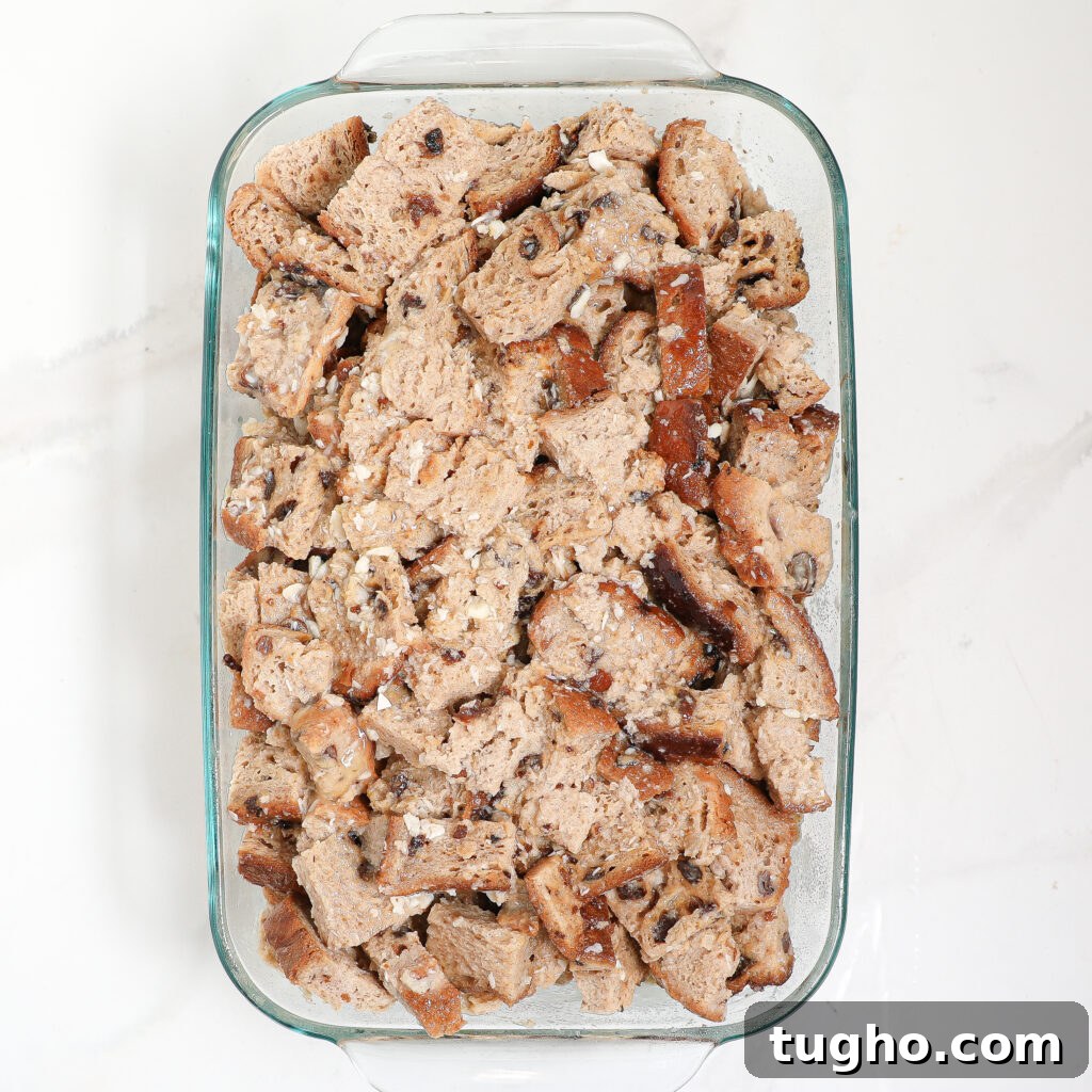 Cinnamon Raisin Bread Pudding Recipe 4 A glass baking dish filled with chunks of cinnamon raisin bread pudding, mixed with raisins or chocolate chips, prepared for baking on a white marble countertop.