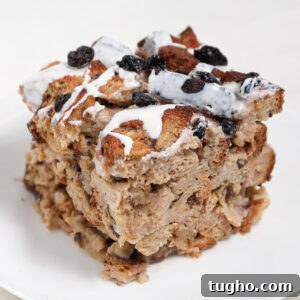 Cinnamon Raisin Bread Pudding Recipe 6 A thick square slice of Cinnamon Raisin Bread Pudding topped with raisins and drizzled with white icing sits on a white plate. The bread is golden brown and appears moist and dense.
