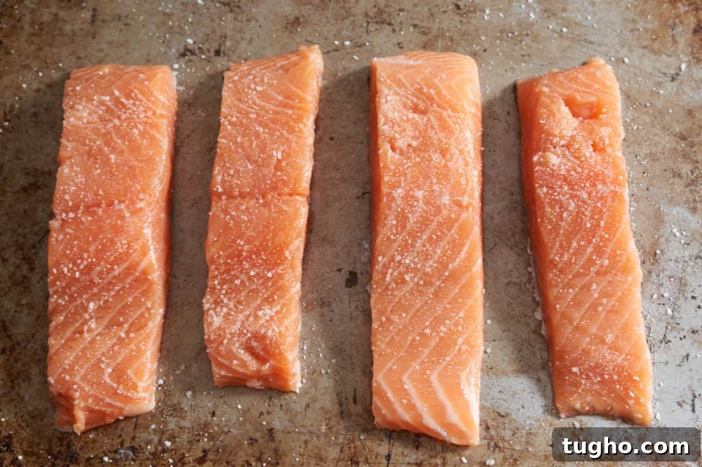 Four filets of raw salmon seasoned with salt on a baking sheet.