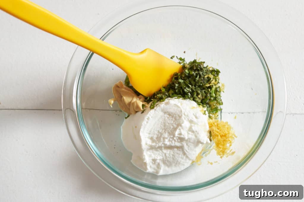A yellow spatula in a glass bowl with yogurt, basil, mustard, and lemon zest.