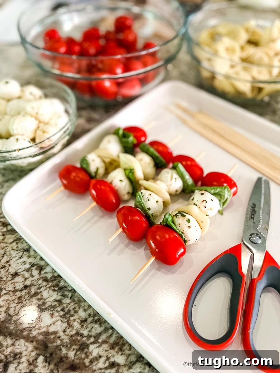 bowls of cherry tomatoes, mozzarella balls, pasta, and basil leaves along with a tray of 4 assembled tortellini appetizers and a pair of scissors on a white serving tray