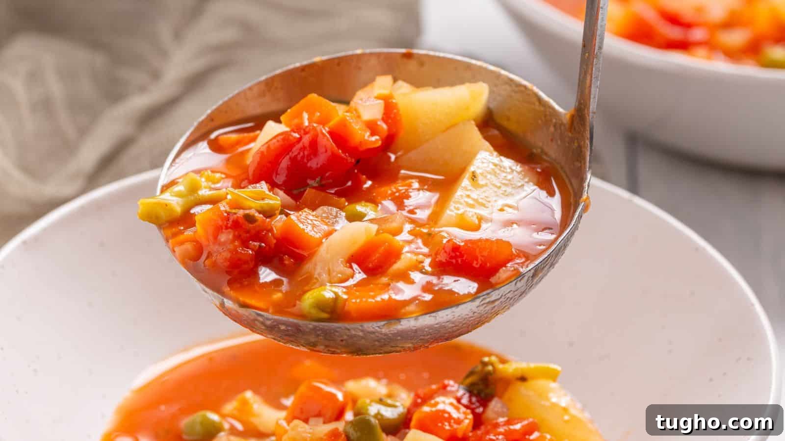 A ladle serving vegetable soup with tomatoes, potatoes, and green peas into a white bowl, with a blurred bowl in the background.