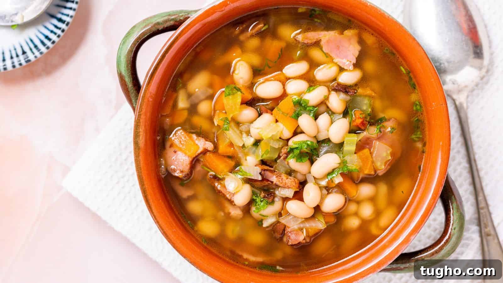 A bowl of bean soup with chunks of ham and diced vegetables, served in a rustic terracotta bowl with a spoon on the side.