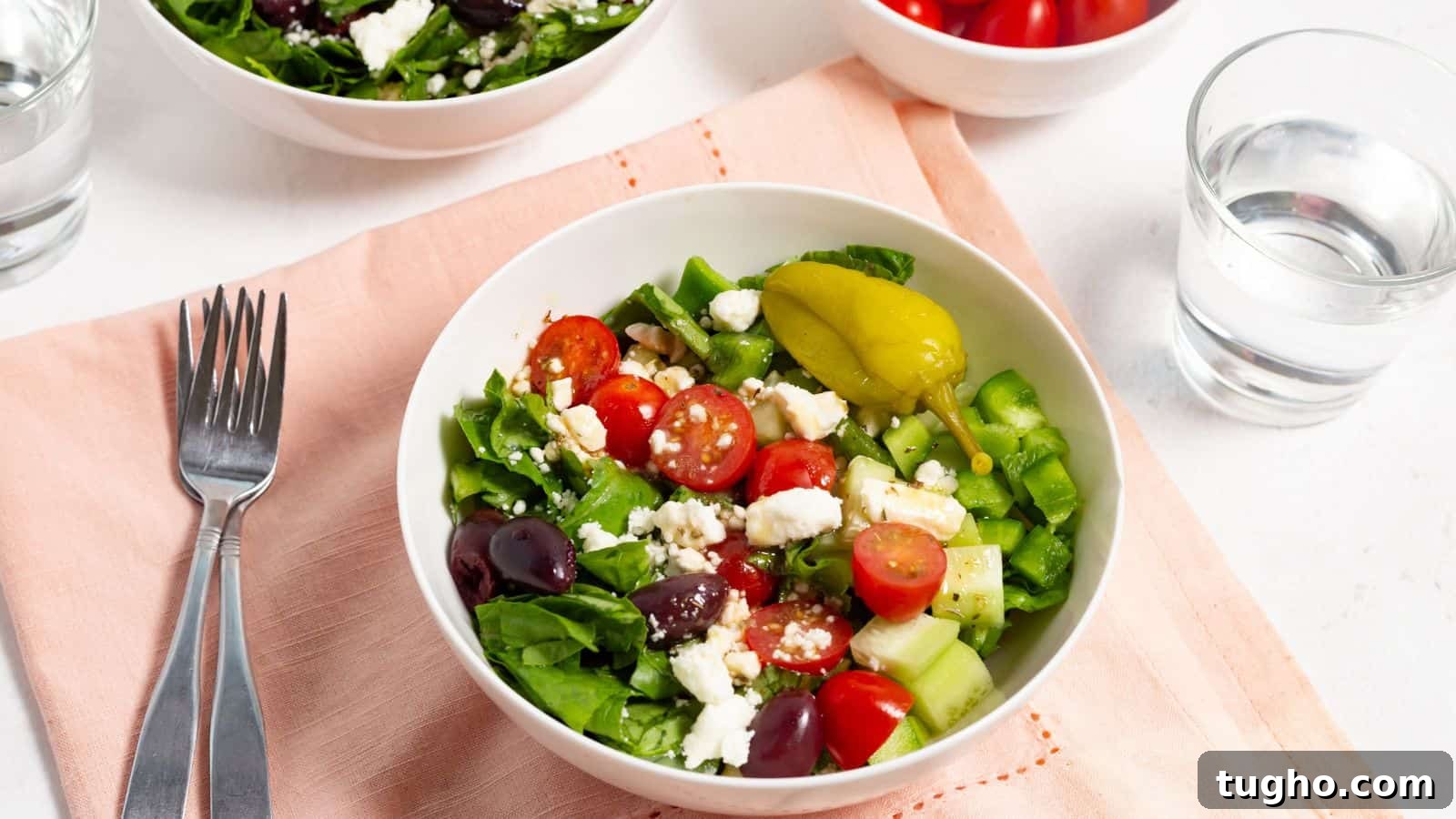 A fresh greek salad with tomatoes, feta cheese, cucumbers, olives, and peppers, served with a fork and a glass of water on the side.