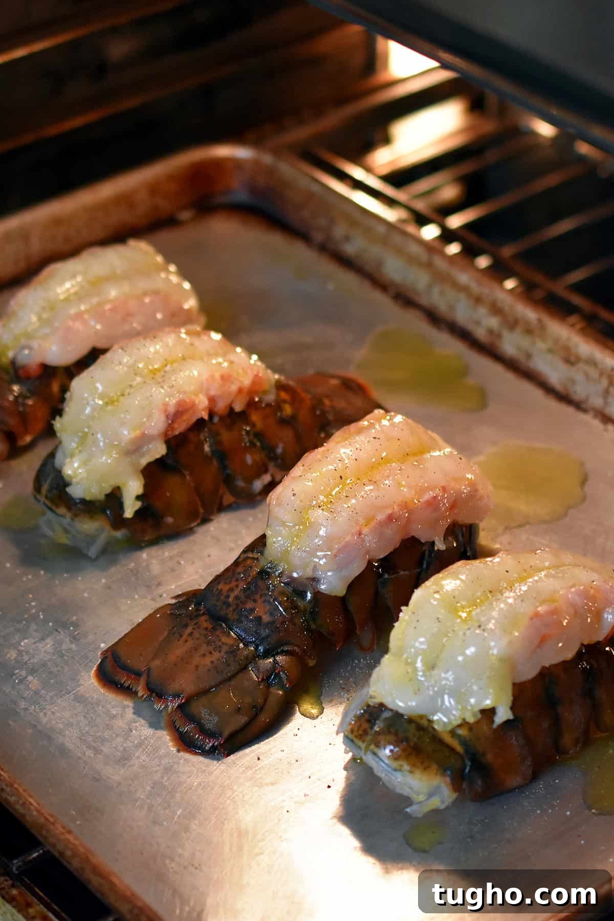 Four butterflied lobster tails on a rimmed baking sheet are baking in the oven.
