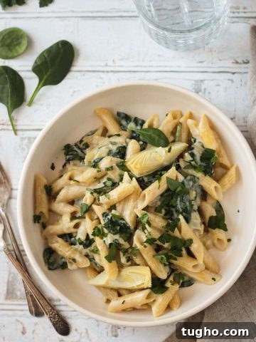 Bowl of vegan spinach artichoke pasta in a white bowl next to two forks.