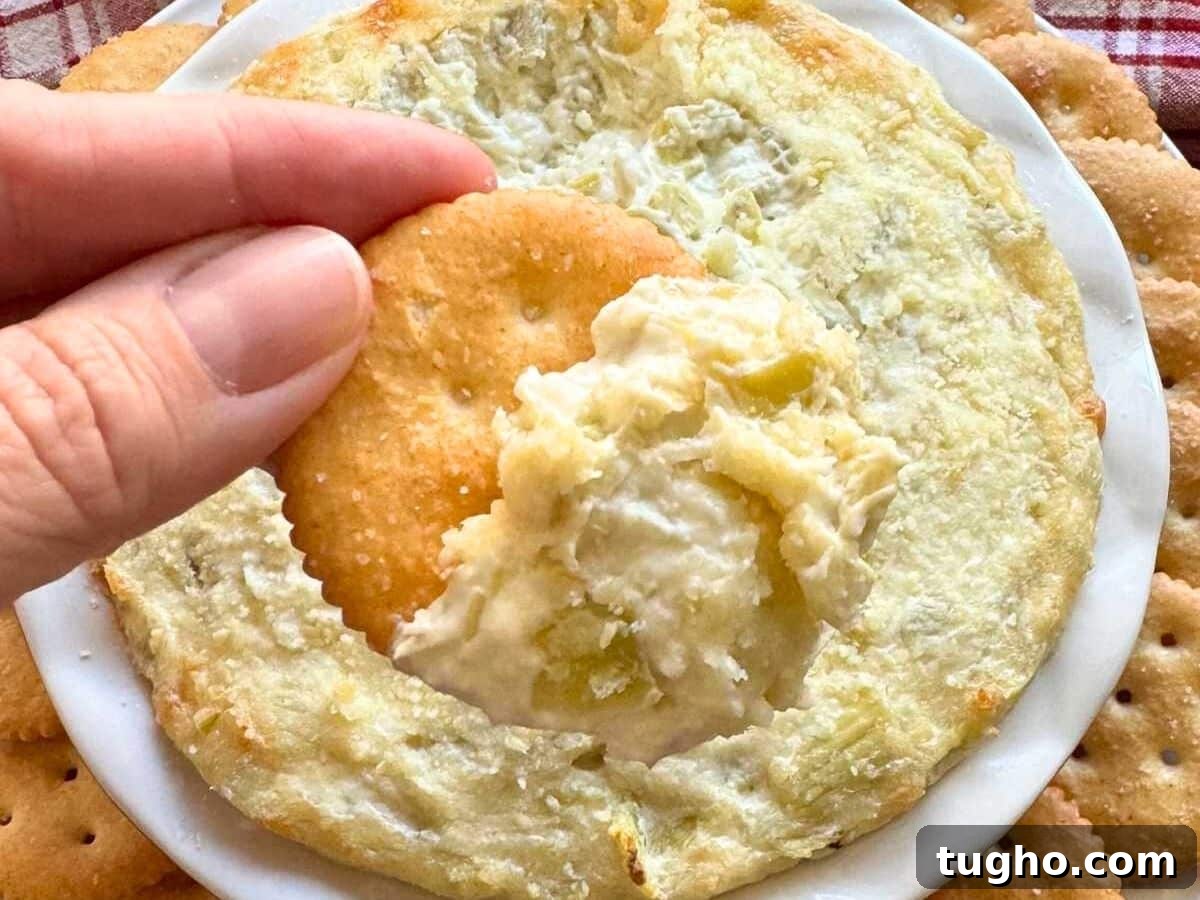 15 Make-Ahead Thanksgiving Recipes to Prep Now 12 Fingers holding a cracker with artichoke dip on it over a bowl with more of it.