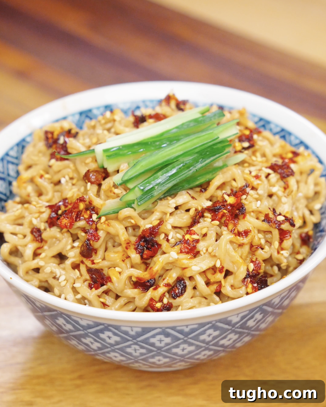Bowl of creamy sesame noodles topped with chili oil, sesame seeds, and sliced cucumbers.