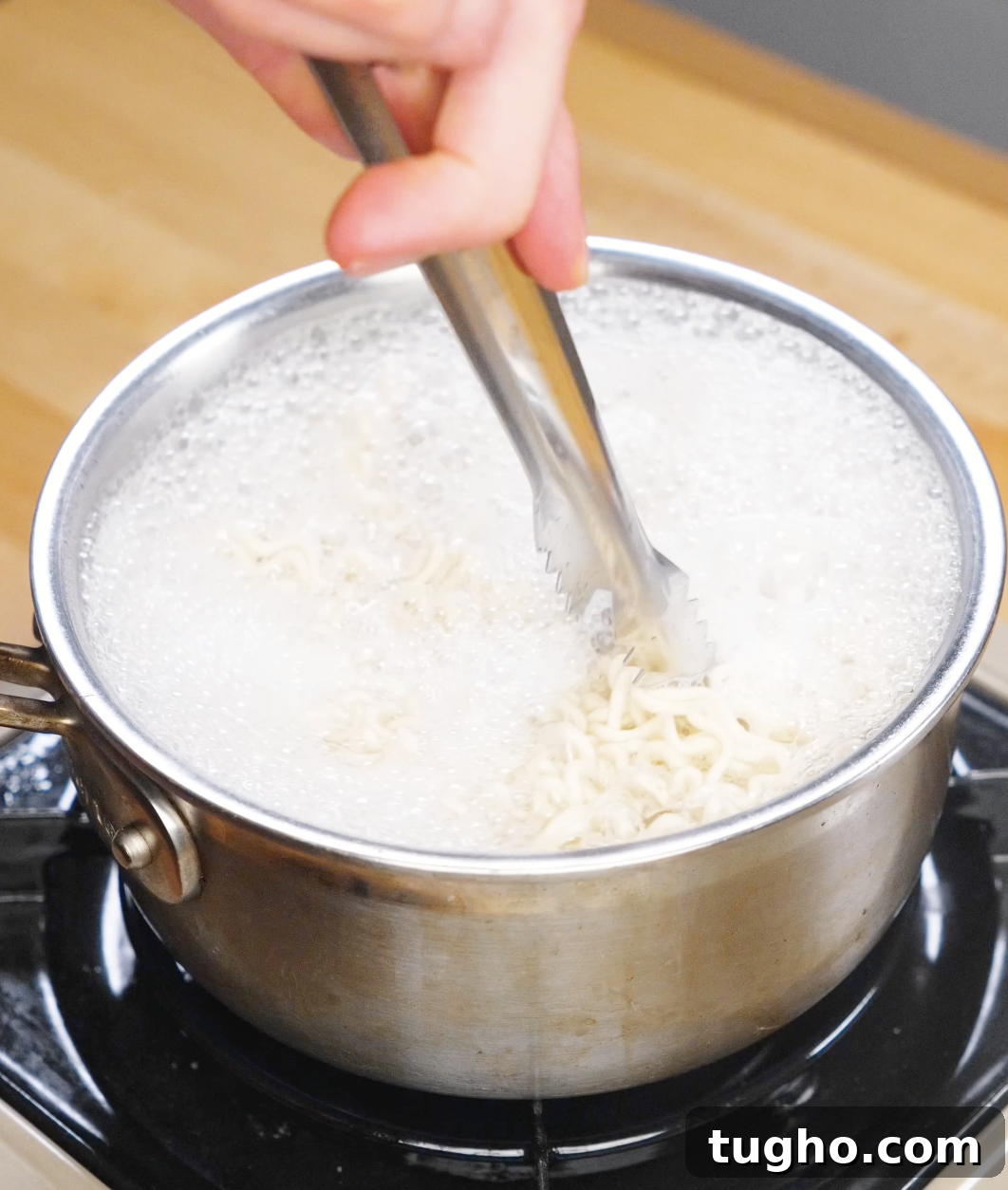 Creamy Sesame Noodles with Garlic Soy Sauce 4 Ramen noodles cooking in a pot of boiling water on the stovetop with tongs stirring.