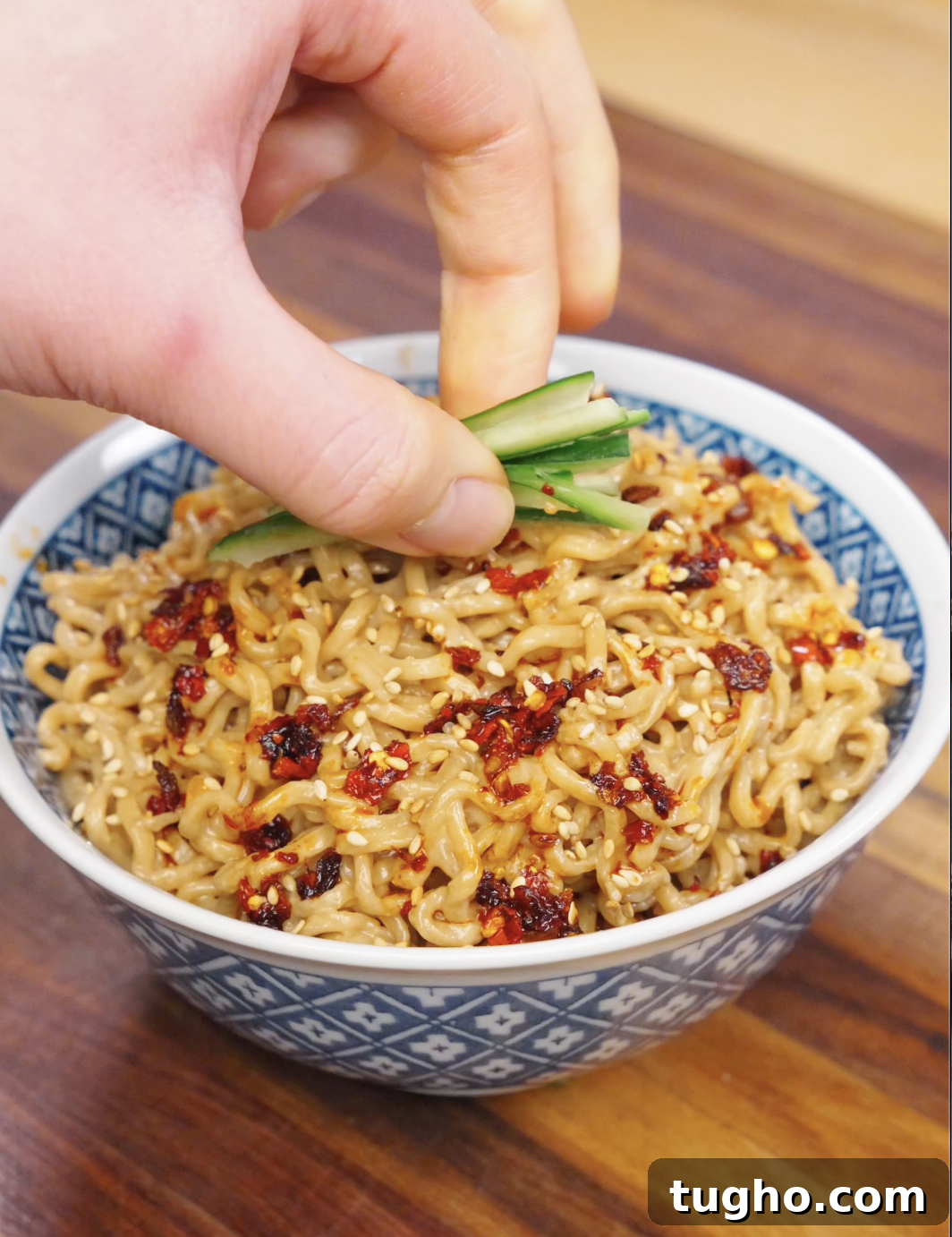 Creamy Sesame Noodles with Garlic Soy Sauce 8 Hand placing sliced cucumber garnish on top of creamy sesame noodles with chili oil and sesame seeds.