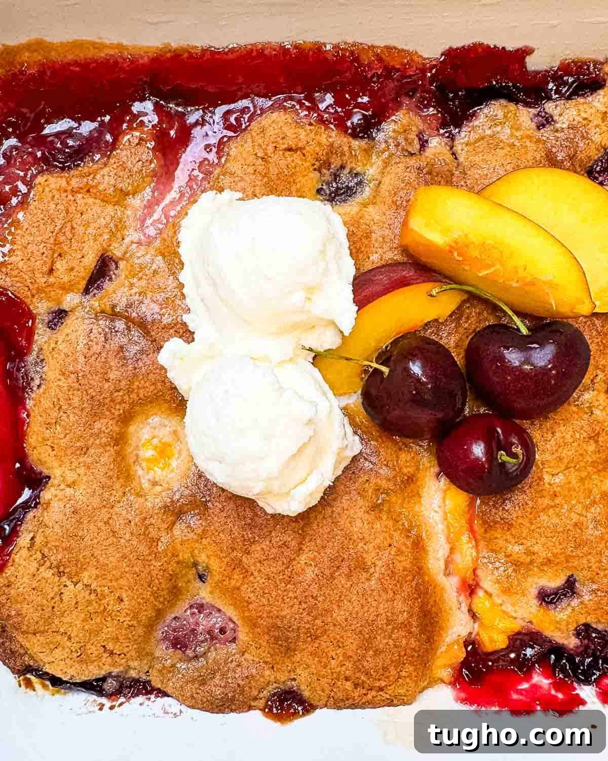 Homemade Cherry and Peach Cobbler Recipe 12 The peach cherry cobbler in a baking dish after it is done baking, topped with ice cream and fresh cherries and peaches for garnish.