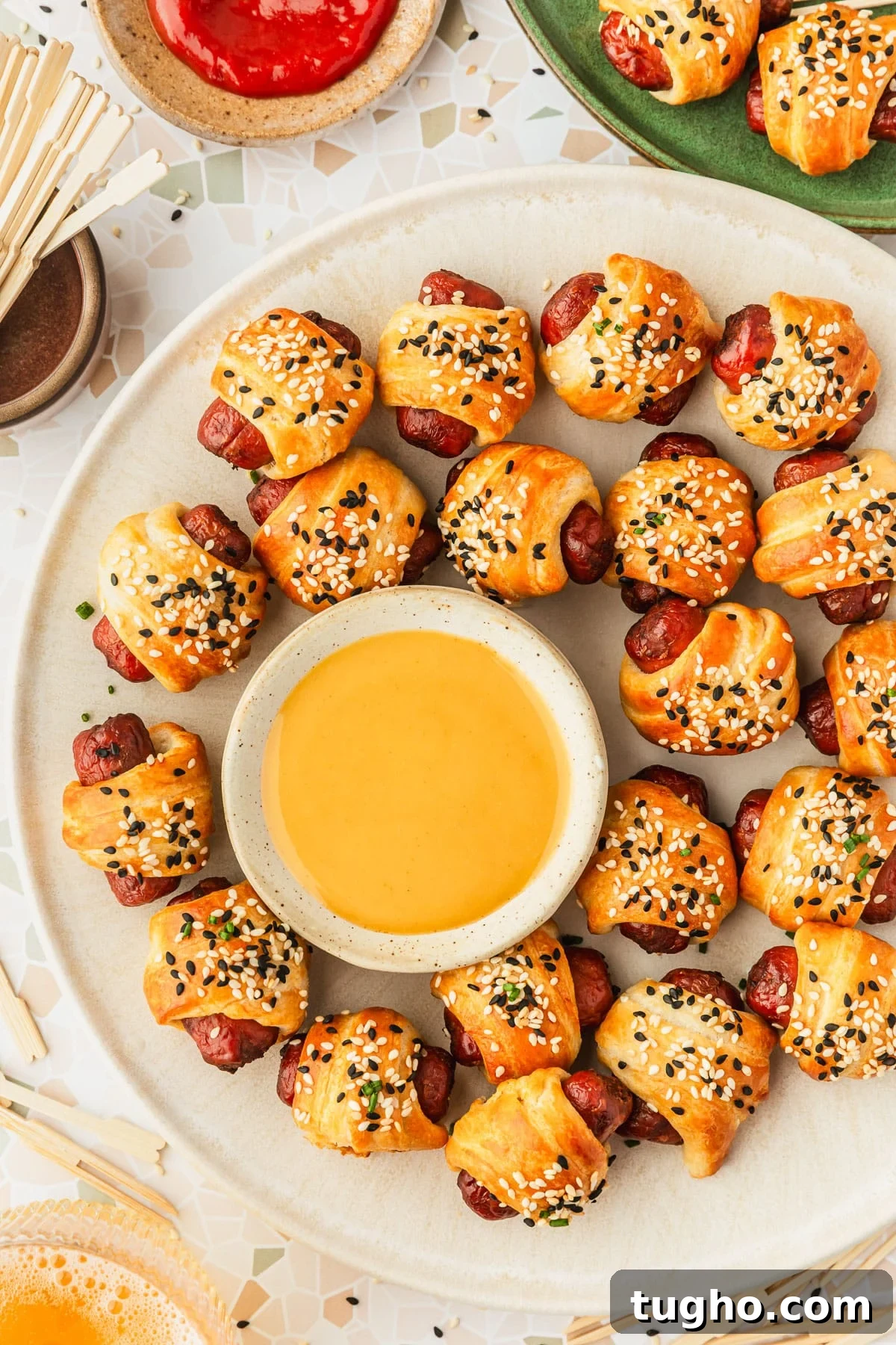 3-Ingredient Air Fryer Pigs in a Blanket Recipe 10 A white platter of air fryer pigs in a blanket on a white terrazzo counter next to a green plate of pigs in a blanket, glass of beer, brown bowl of toothpicks, and brown bowl of ketchup.