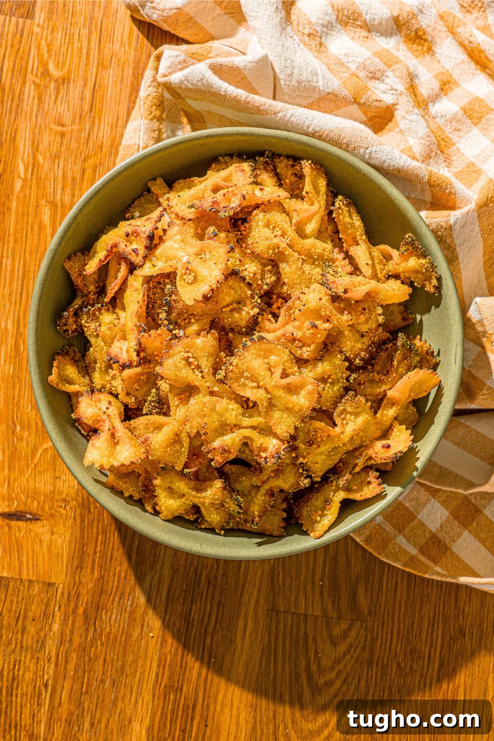 Crispy Baked Pasta Chips Recipe 2 Air fryer pasta chips in a green bowl that is on top of a wood counter with a yellow and white check towel around the front right side of the bowl.