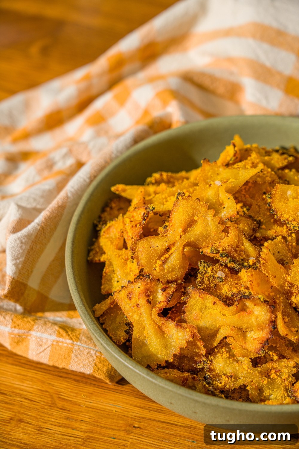 Crispy Baked Pasta Chips Recipe 10 A green bowl with air fried bowtie pasta chips tossed in parmesan and mac and cheese powder. The bowl is on a wooden counter with a yellow striped napkin.