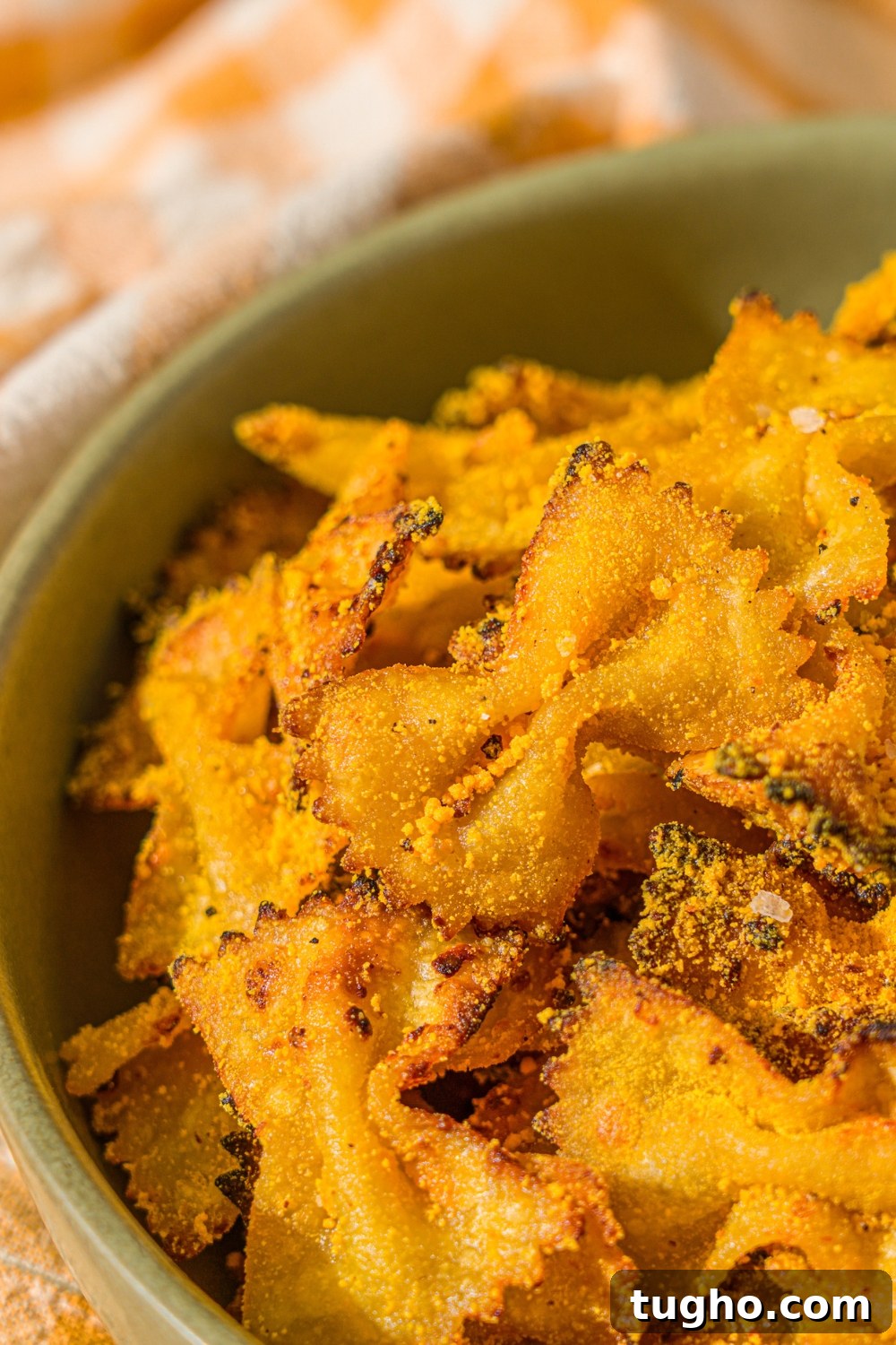 Crispy Baked Pasta Chips Recipe 11 A green bowl with air fried bowtie pasta chips tossed in parmesan and mac and cheese powder. The bowl is on a wooden counter with a yellow striped napkin.