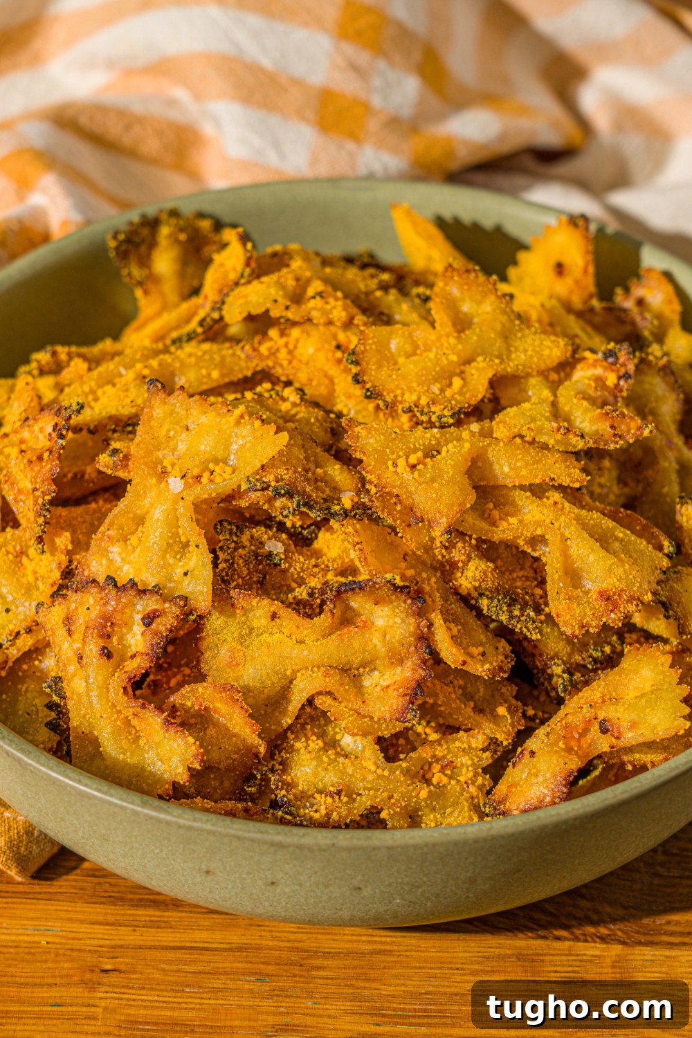 Crispy Baked Pasta Chips Recipe 3 A green bowl with air fried bowtie pasta chips tossed in parmesan and mac and cheese powder. The bowl is on a wooden counter with a yellow striped napkin.
