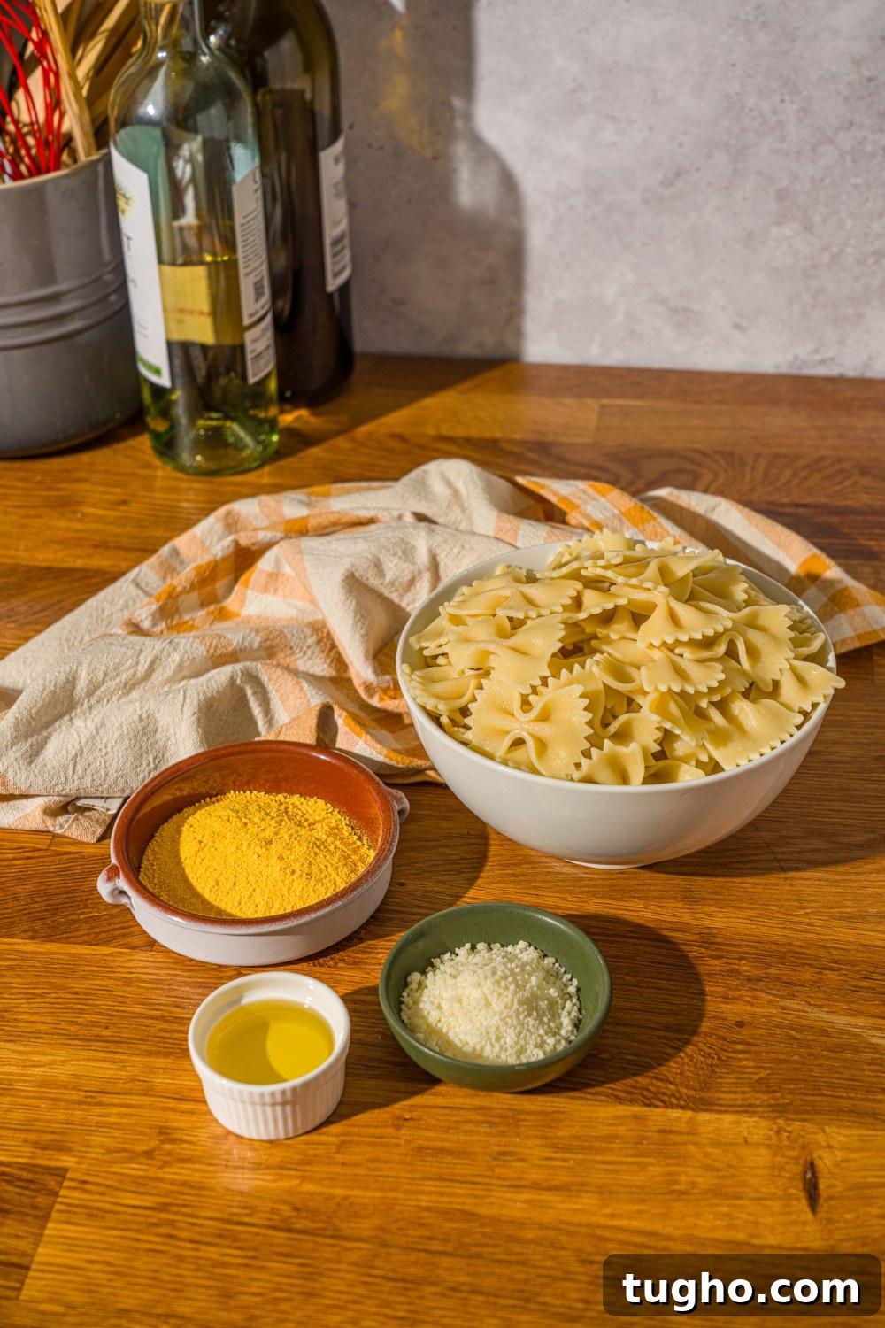 Crispy Baked Pasta Chips Recipe 4 A wooden counter with ingredients to make pasta chips including a bowl of bowtie pasta, oil, parmesan cheese, mac and cheese powder, and a yellow striped napkin.