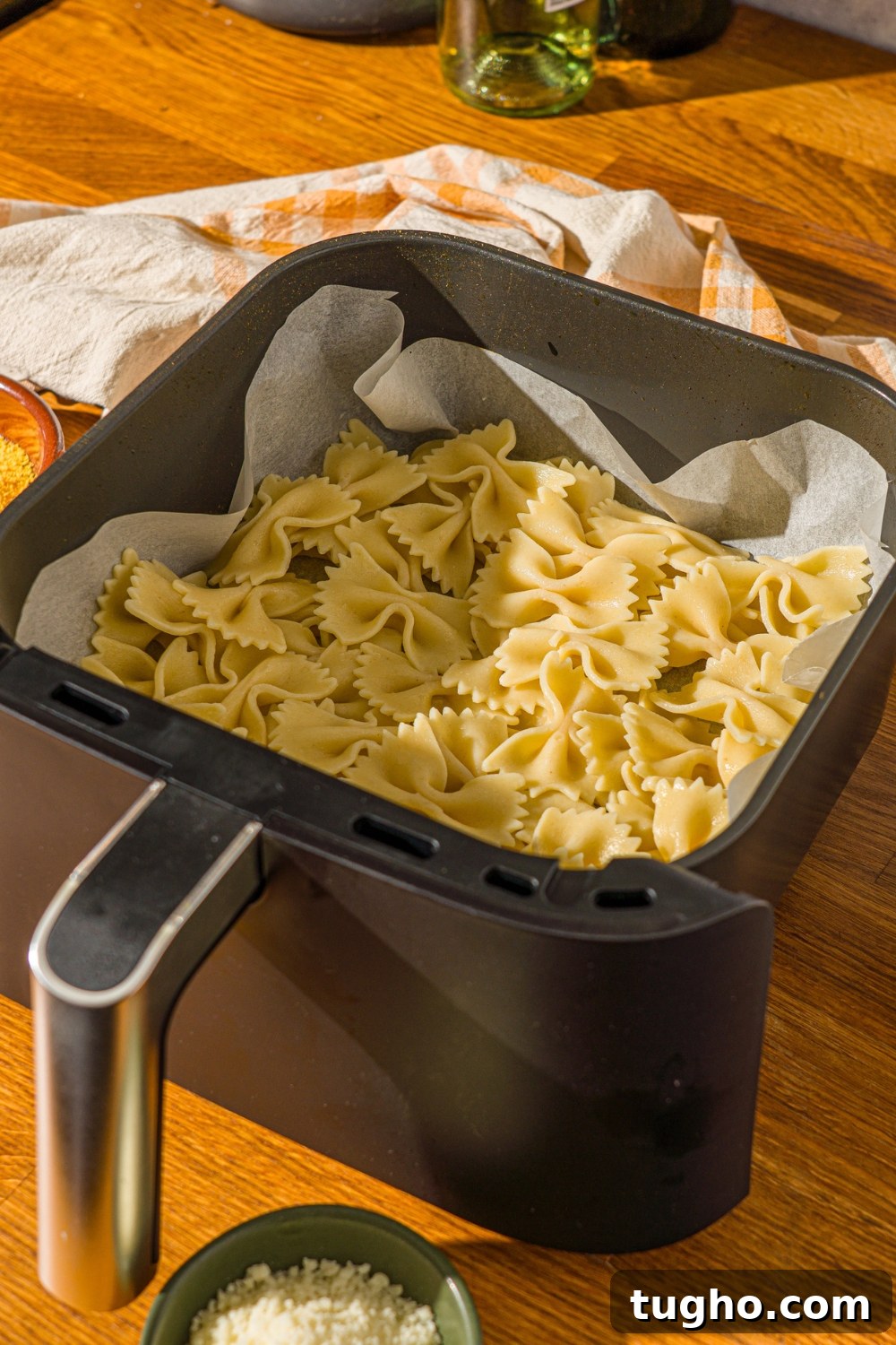 Crispy Baked Pasta Chips Recipe 5 An air fryer basket lined with parchment paper with cooked bowtie pasta. The basket is on a wooden counter with a yellow striped napkin.