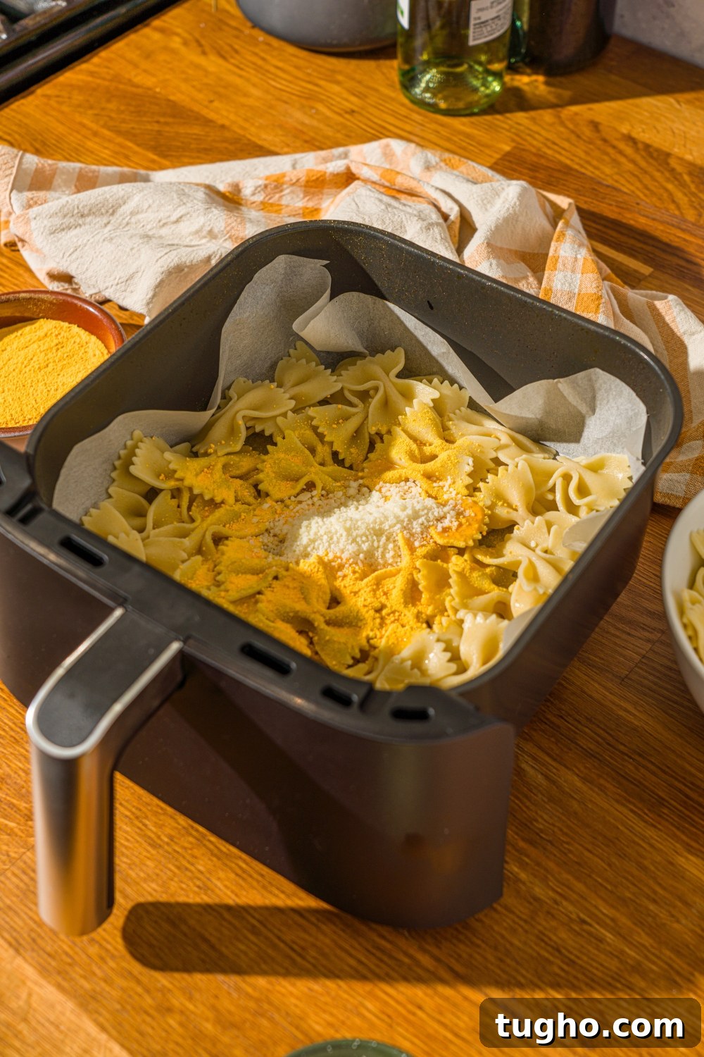 Crispy Baked Pasta Chips Recipe 6 An air fryer lined with parchment paper with cooked bowtie pasta coated in oil and topped with parmesan cheese and mac and cheese powder. The air fryer is on a wooden counter with a yellow striped napkin.