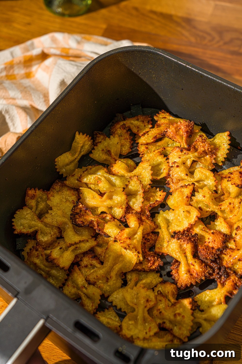 Crispy Baked Pasta Chips Recipe 7 An air fryer basket with air fried bowtie pasta chips seasoned in cheese. The basket is on a wooden counter with a yellow striped napkin.