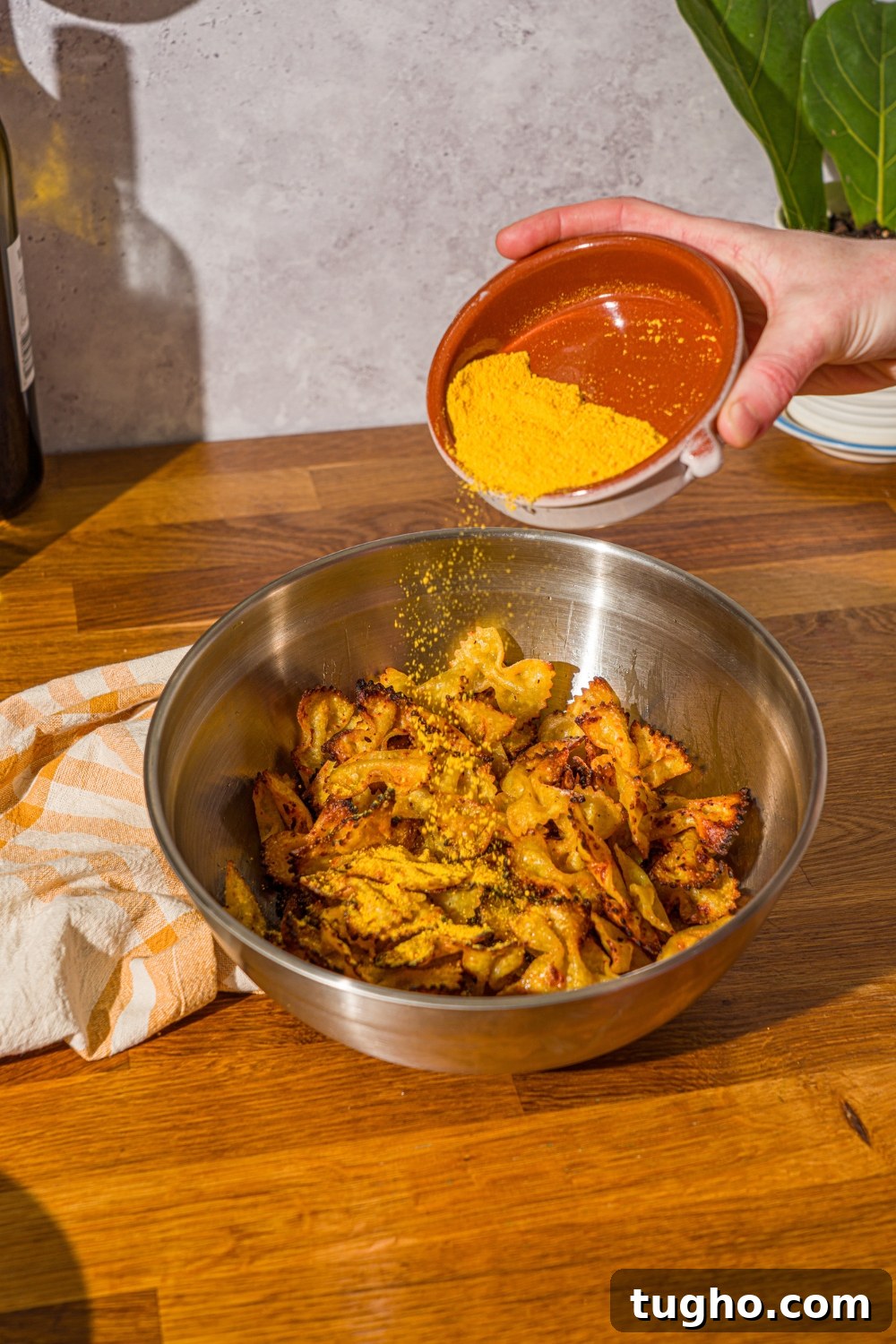 Crispy Baked Pasta Chips Recipe 8 A mixing bowl with air fried bowtie pasta chips with a bowl of mac and cheese powder being poured over the chips. The bowl is on a wooden counter with a yellow striped napkin.