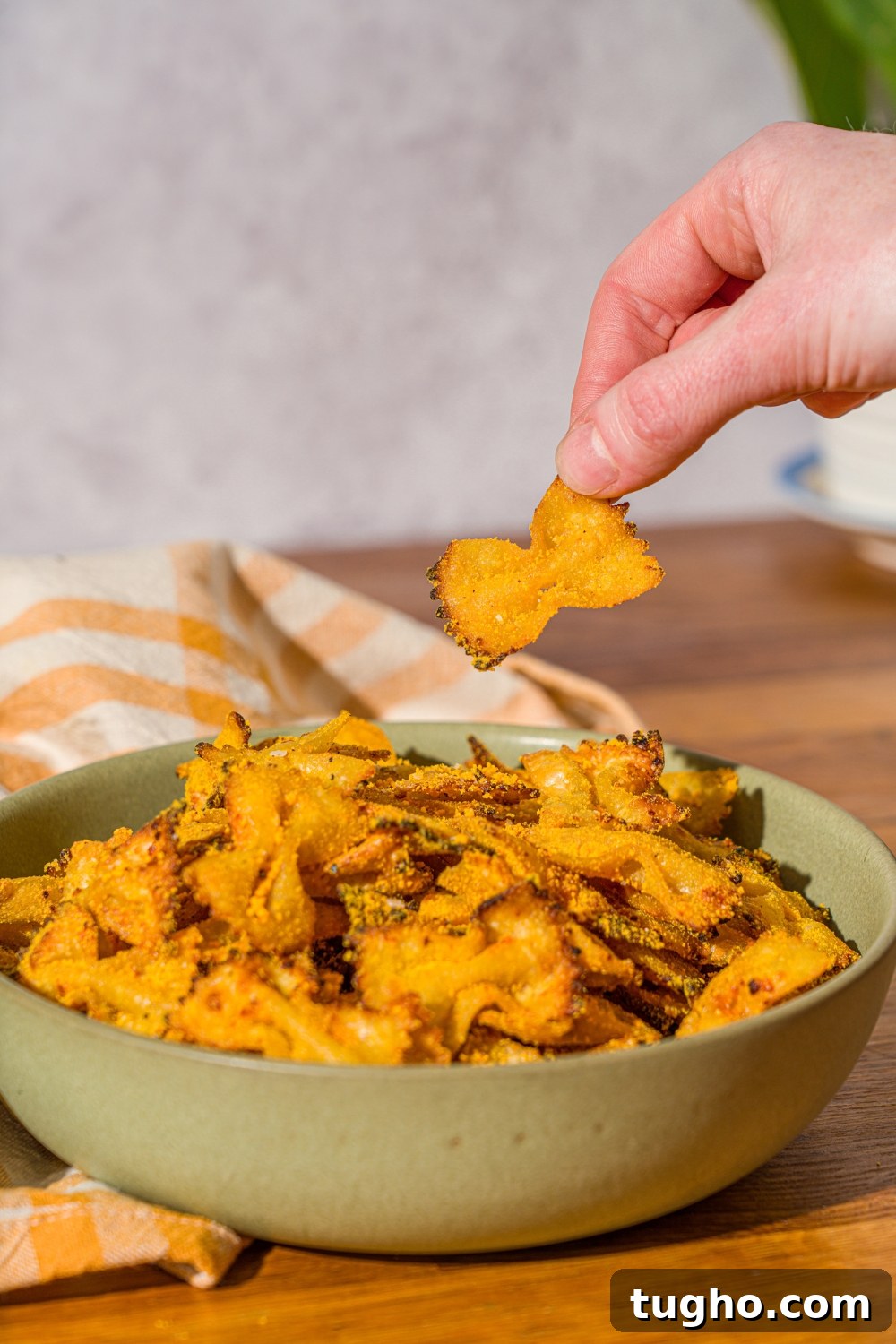 Crispy Baked Pasta Chips Recipe 9 A bowl of bowtie pasta chips seasoned with cheese. There is a hand taking a pasta chip from the bowl. The bowl is on a wooden counter with a yellow striped napkin.