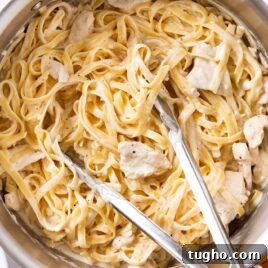 Large metal stockpot filled with fettuccine alfredo with pieces of chicken resting on top along with tongs all on white marble surface.