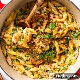 Large Dutch oven filled with tuna noodle casserole topped with melted cheese, chopped parsley, and browned bread crumbs with wooden spoon resting in bowl.