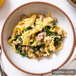 White bowl with brown rim filled with creamy sausage pasta and greens all on white surface.