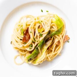 White plate filled with spaghetti noodles with brown butter sauce on top with shaved asparagus and bacon and chive pieces