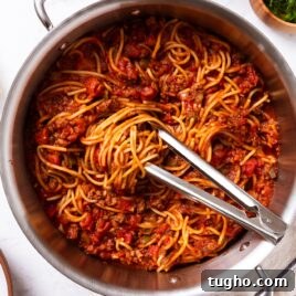 Large metal stockpot filled with meat sauce with spaghetti with tongs sitting on top with swirl of noodles all on marble surface with towel, green herbs, and Parmesan cheese.