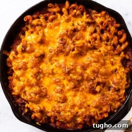 Black cast iron skillet with homemade goulash baked inside sprinkled with cheddar cheese on top with towel covering handle of skillet.