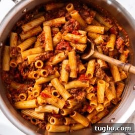 White marble surface with metal stockpot filled with cooked bolognese sauce tossed with pasta with wooden spoon resting on bottom of pan.